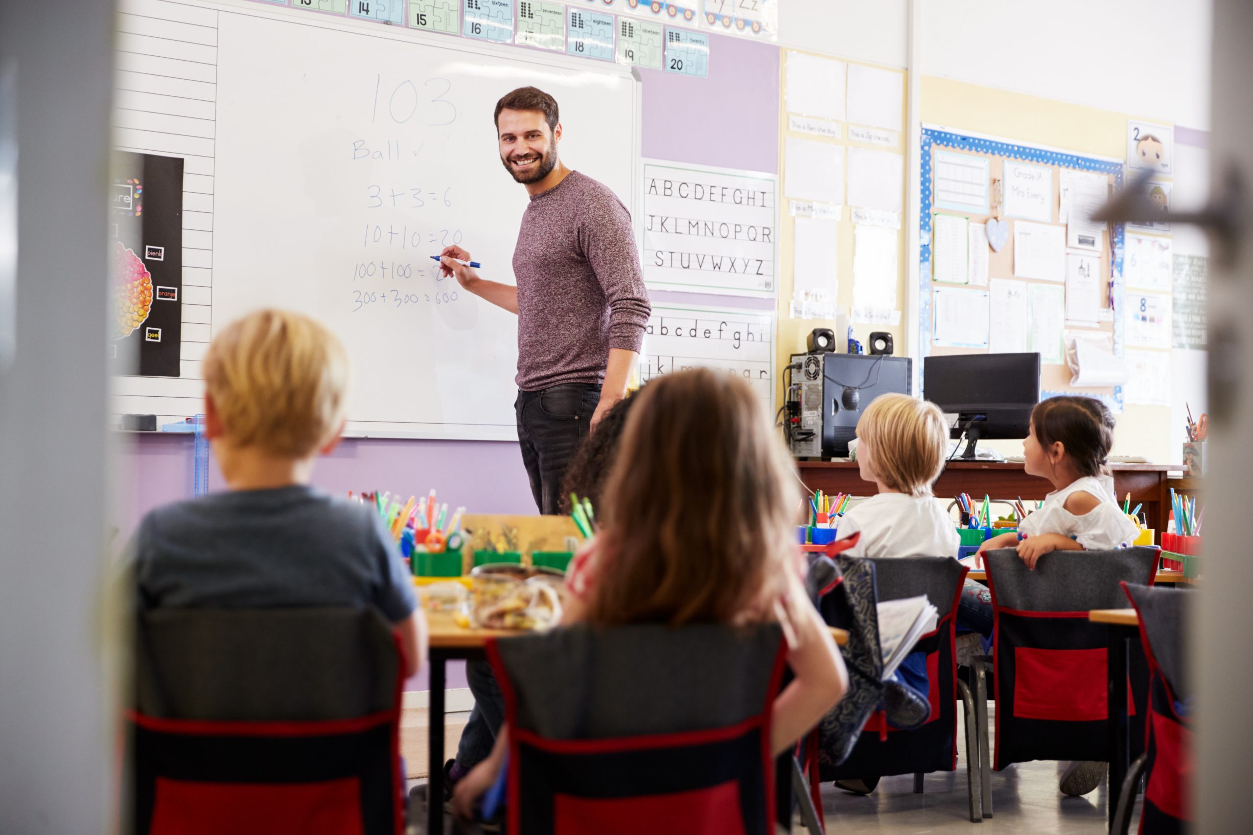 Male,Teacher,Standing,At,Whiteboard,Teaching,Maths,Lesson,To,Elementary